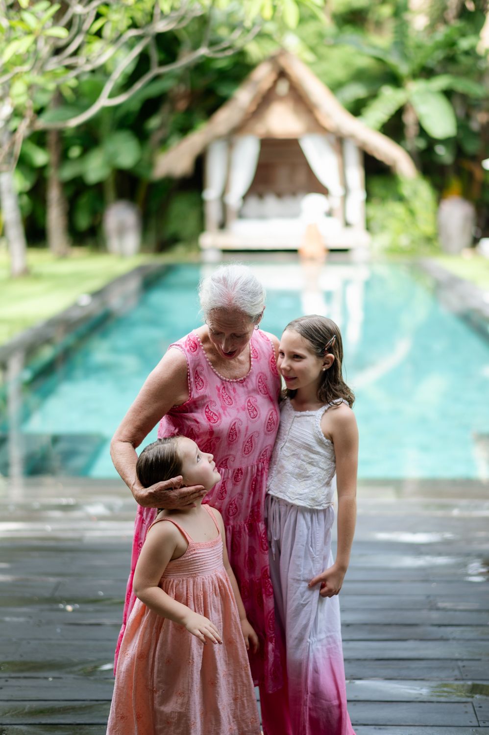 Grandmother in pink dress tenderly interacting with two young granddaughters beside a tropical pool and garden gazebo.