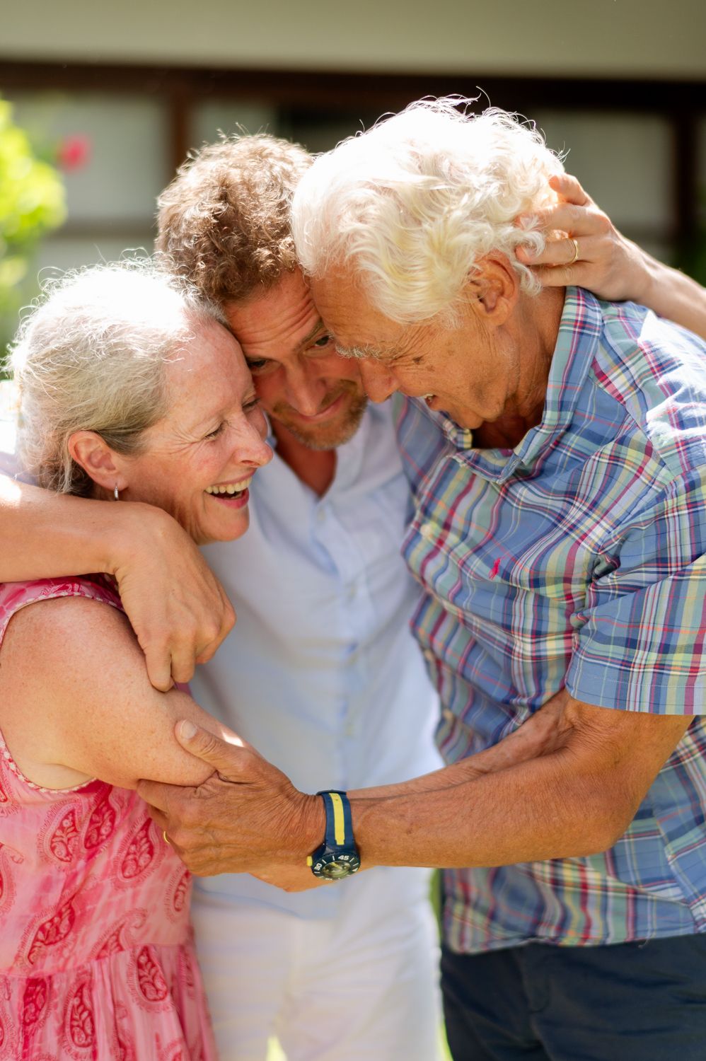 Three elderly people sharing a warm group hug outdoors, smiling and laughing together on a sunny day.