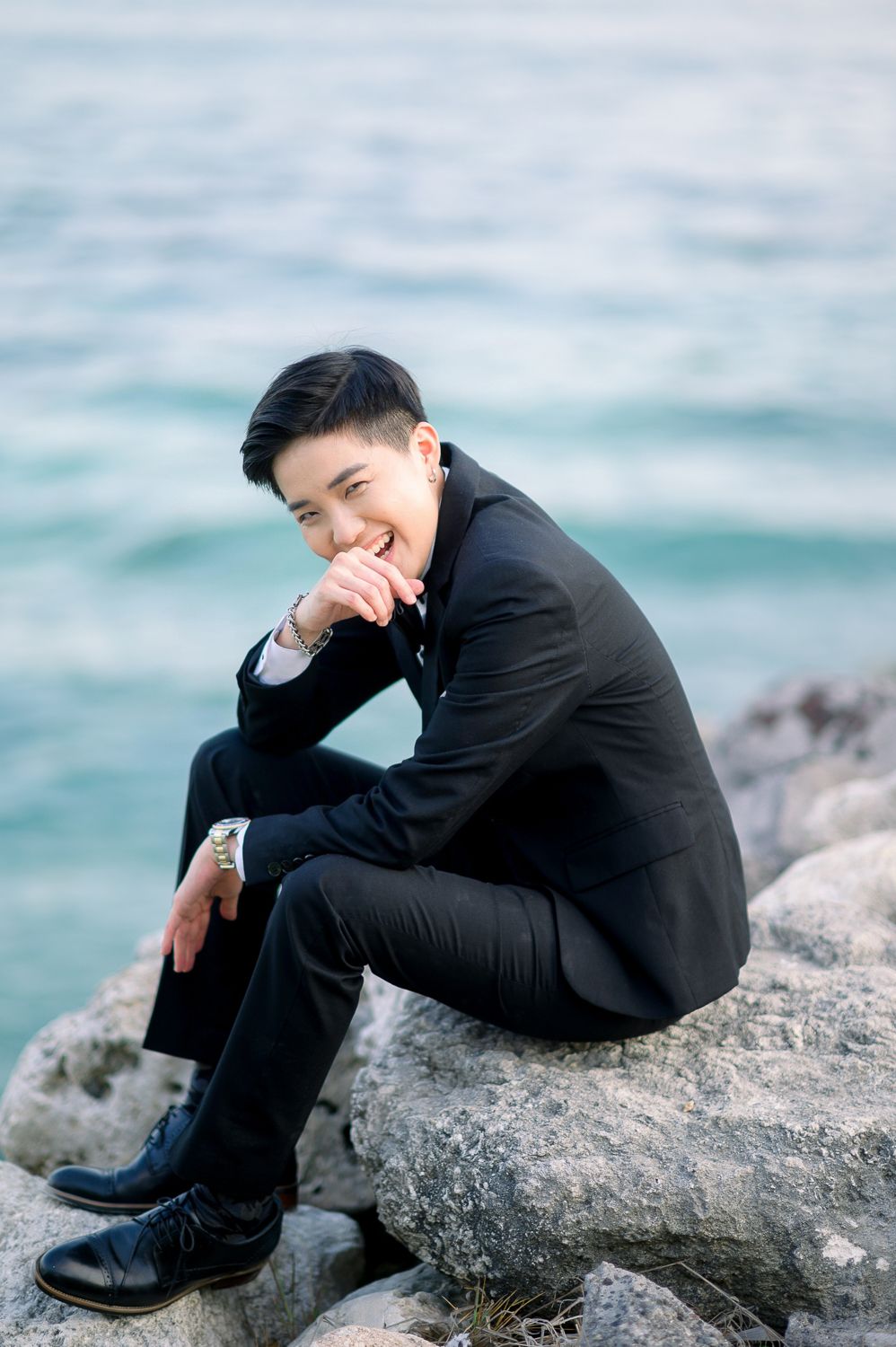 Young man in black suit sitting on rocks by the ocean, smiling confidently in a professional portrait photo.