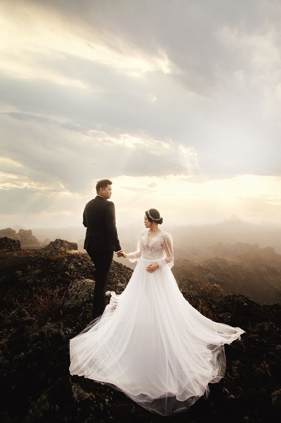 A bride in a flowing white gown and groom in black suit hold hands on a rocky mountaintop at sunset.