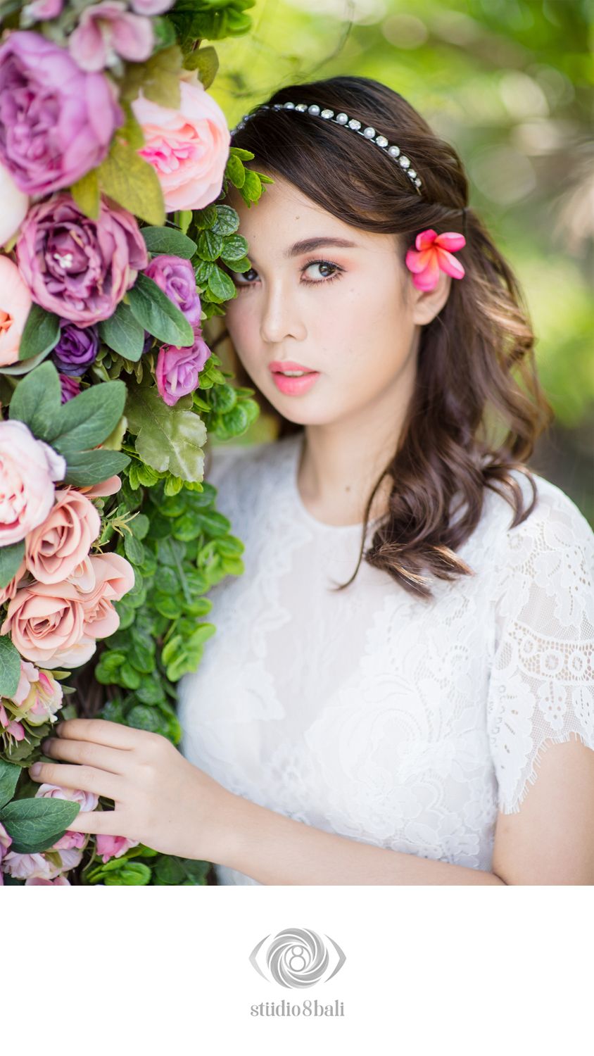 Young bride in white lace dress with floral headband poses beside colorful pink and purple roses in a romantic bridal portrait.