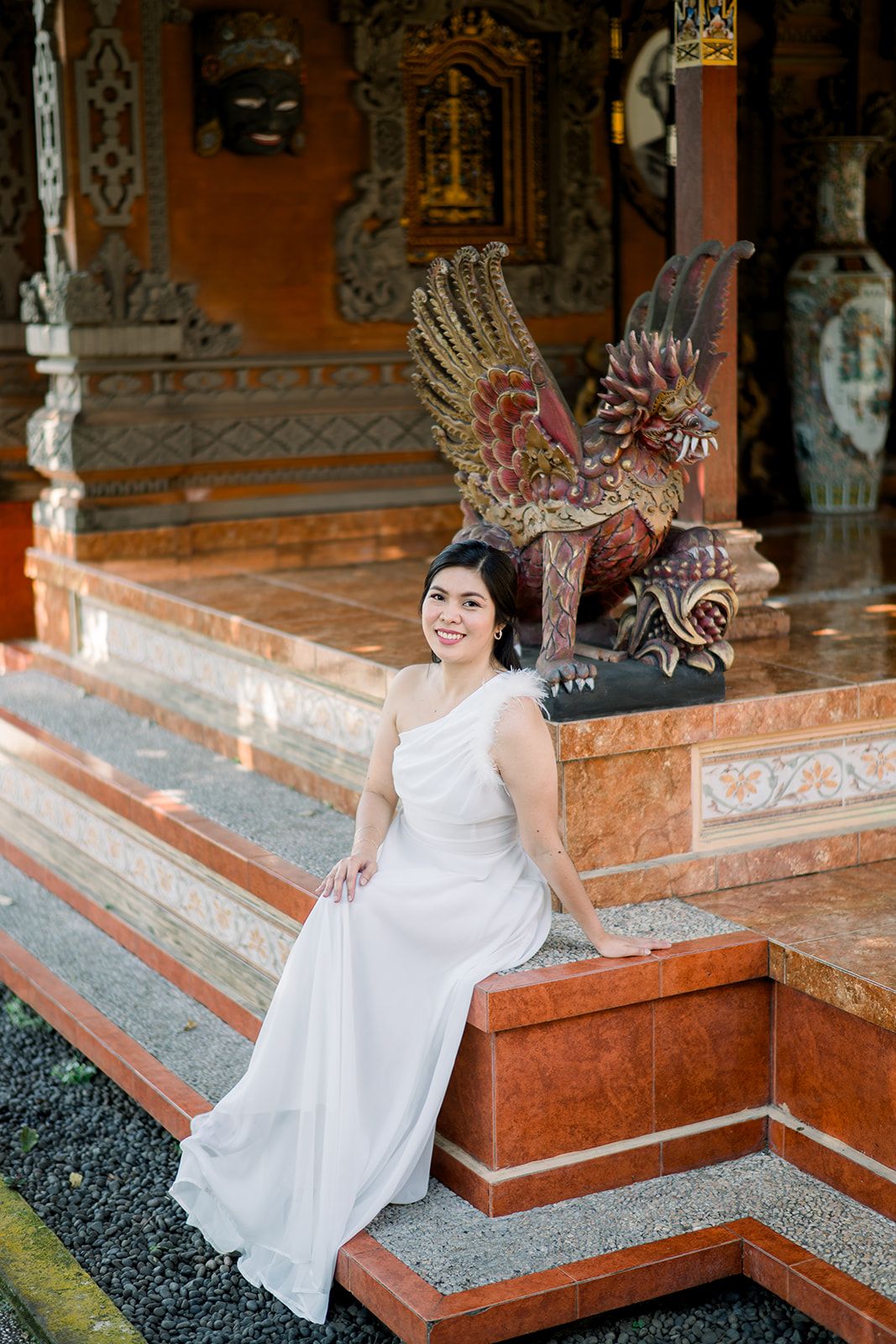 Smiling Asian woman in white dress sitting on temple steps beside ornate Garuda statue at Balinese Hindu temple.