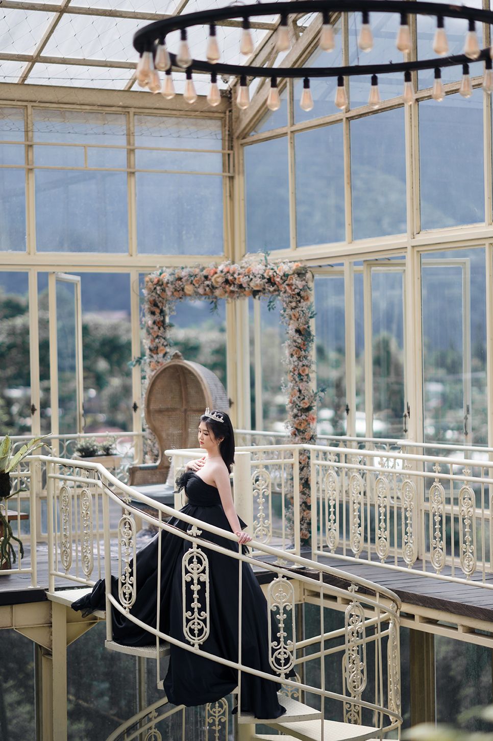 Woman in black gown wearing tiara stands on ornate staircase in glass greenhouse with floral arch and mountain views.
