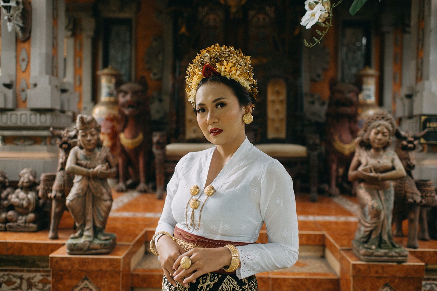 Woman in traditional Balinese attire with gold headdress poses gracefully among ornate stone sculptures at a temple.
