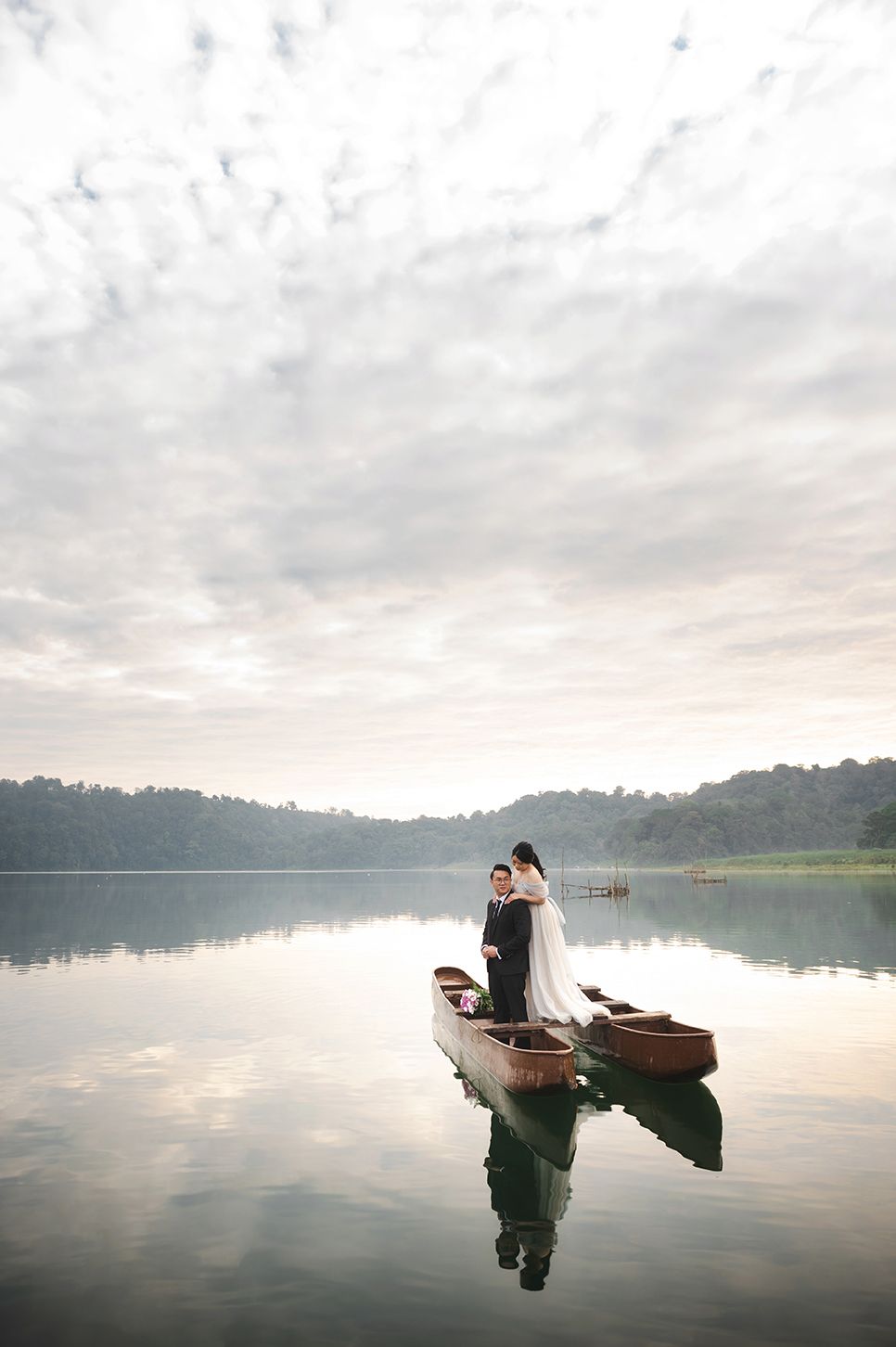 Bride and groom standing on a wooden boat on a calm reflective lake surrounded by misty mountains.