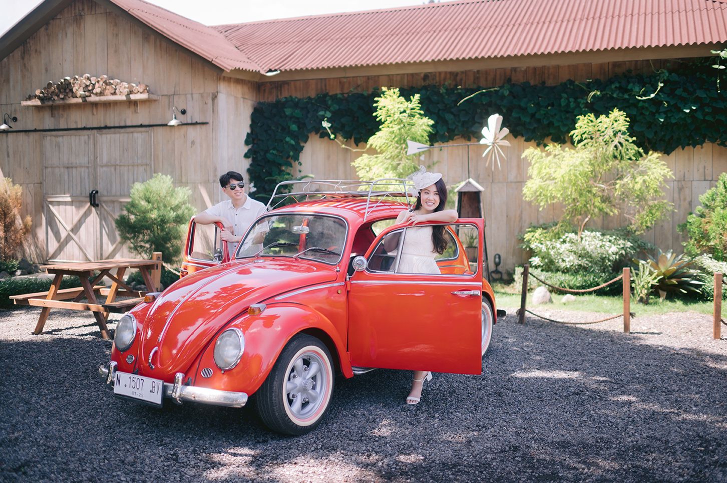 Couple posing with a classic red Volkswagen Beetle convertible outside a rustic barn venue on a sunny day.