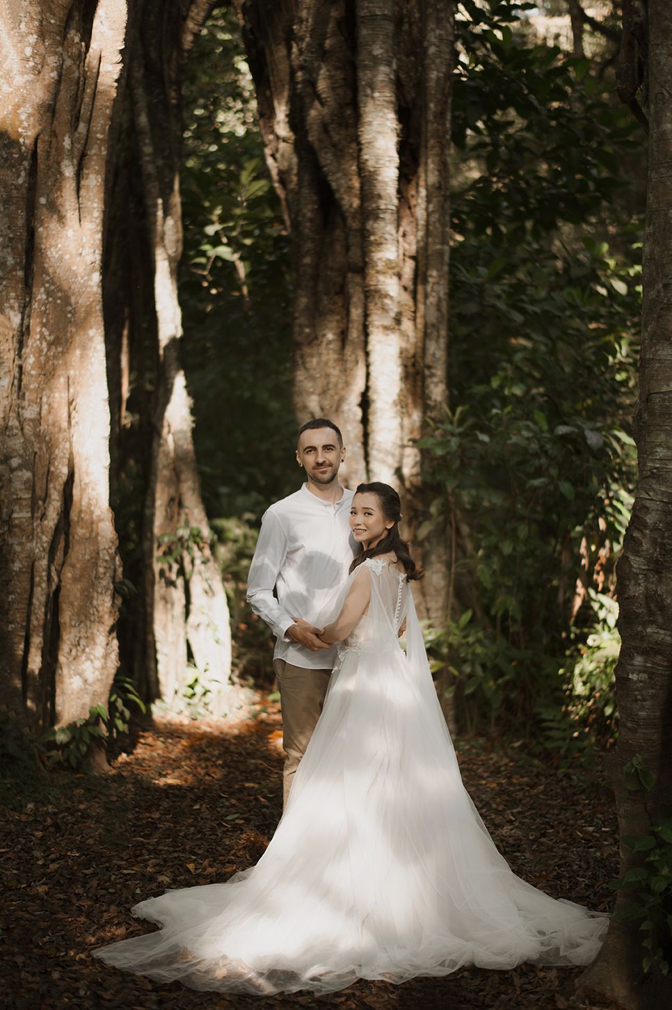 A bride and groom pose romantically in a lush forest, her white gown trailing elegantly on the leaf-covered ground.