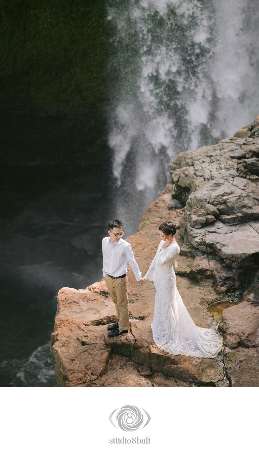 Couple in wedding attire holding hands on rocky ledge before a dramatic waterfall backdrop in Bali.