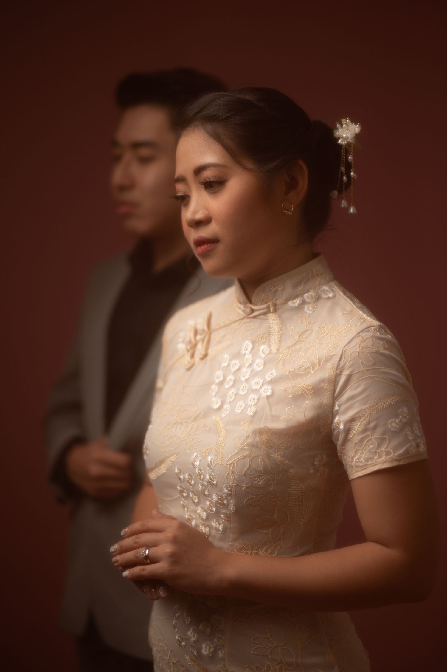 A bride in a white lace qipao cheongsam with floral hair accessories stands before a groom in a gray suit.