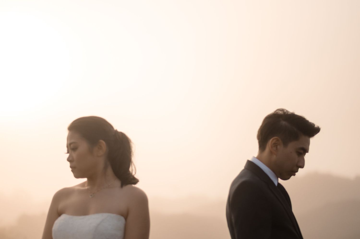 A bride and groom stand apart, looking away from each other, in a moody, hazy outdoor wedding portrait.