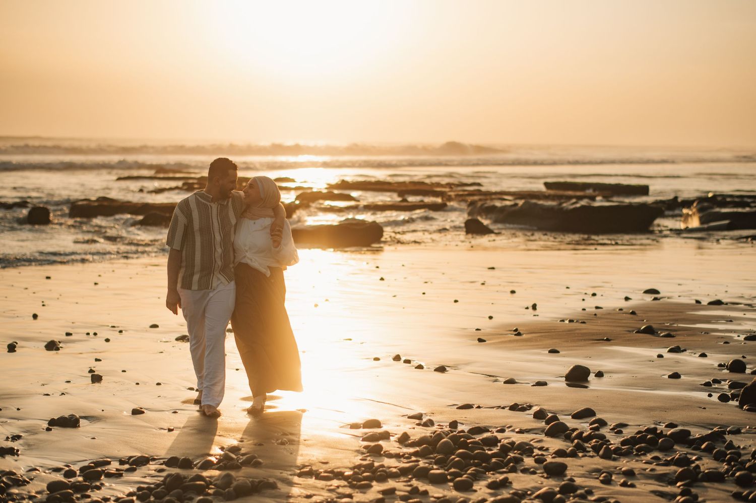 Couple embracing on a rocky beach at golden sunset, warm light reflecting on wet sand and ocean waves.