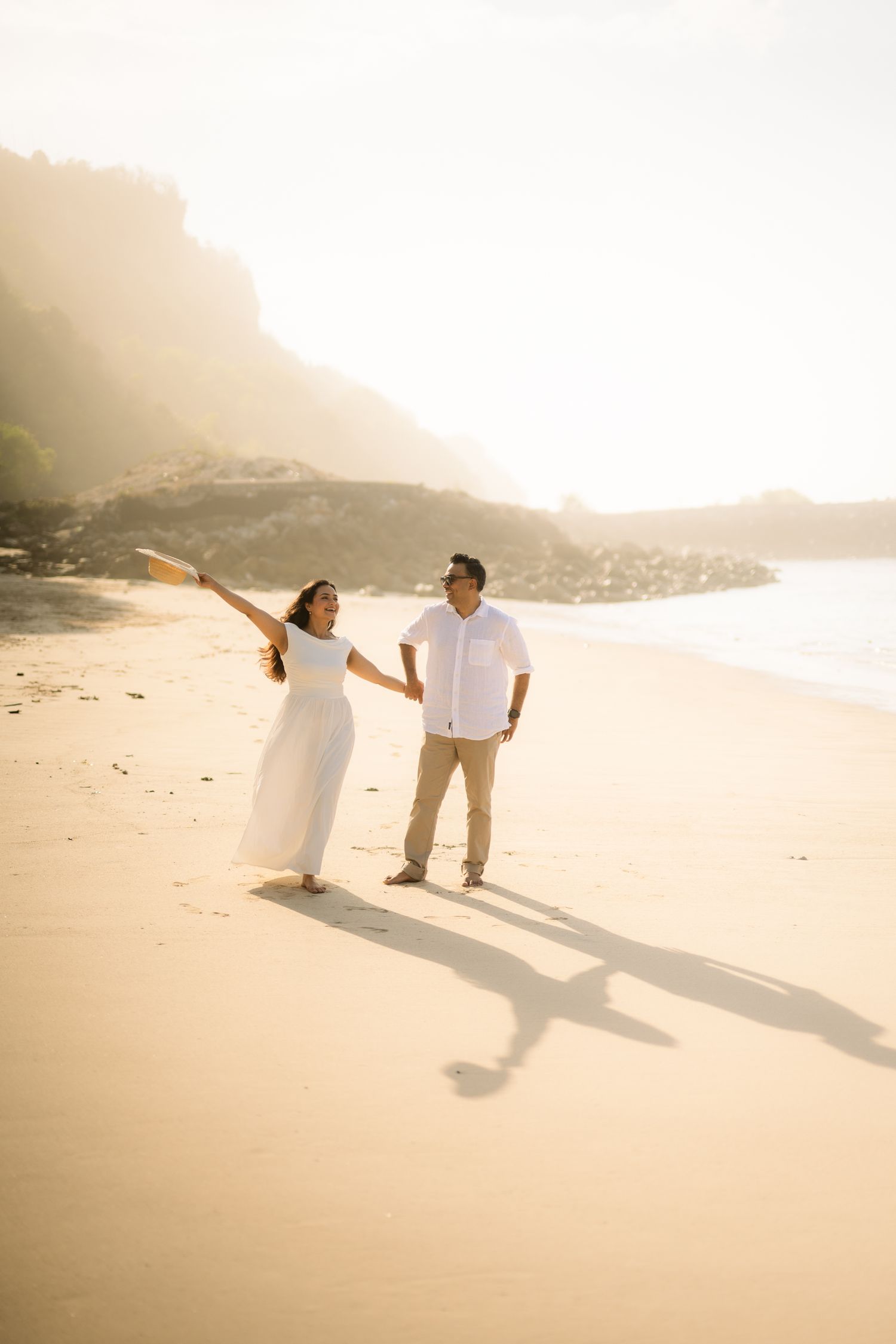 Couple in white outfits holding hands on a sunny beach with rocky cliffs, woman raising her arm joyfully.