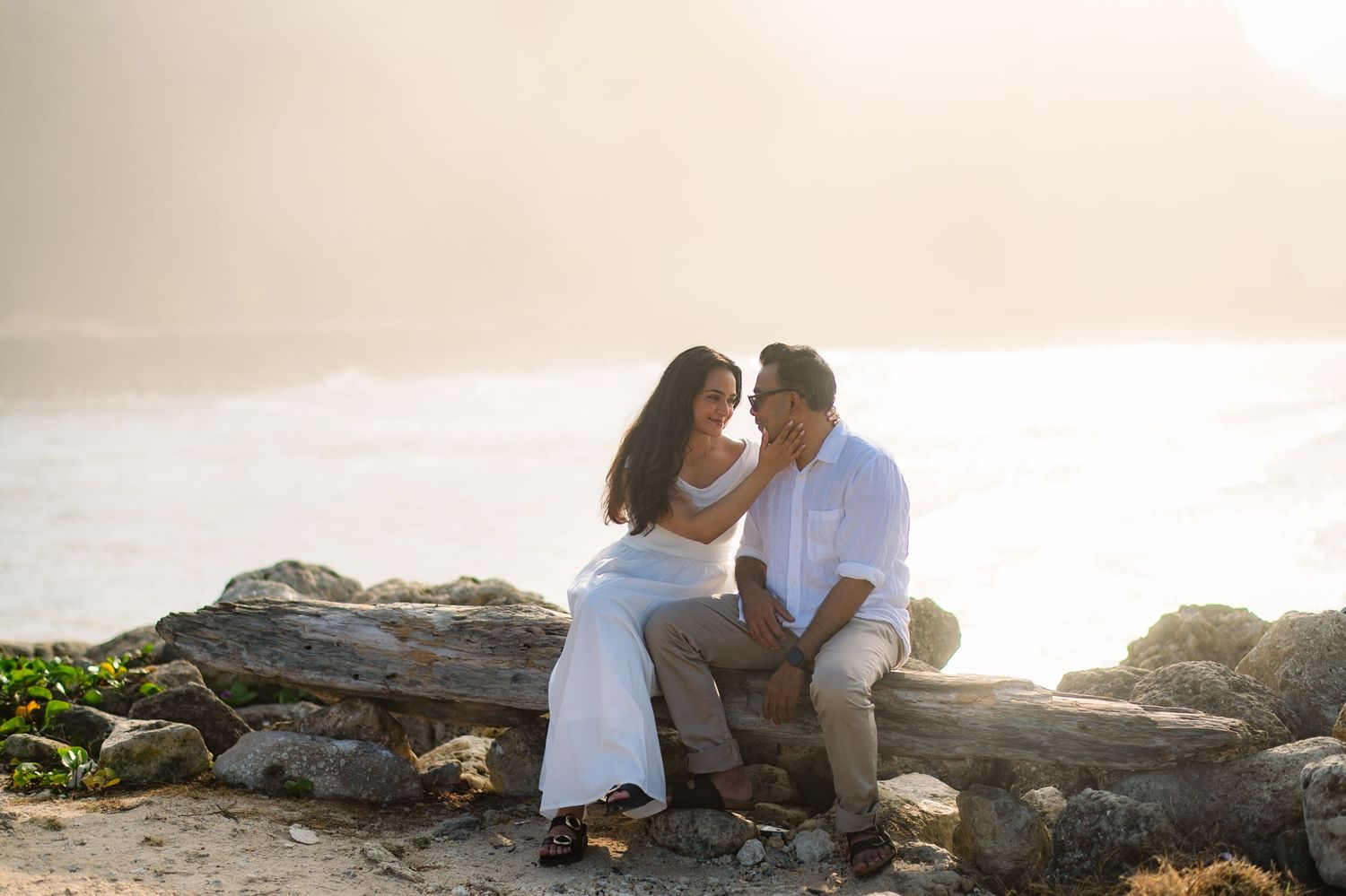 Couple sitting on driftwood by rocky ocean shore in golden sunset light, sharing an intimate moment.