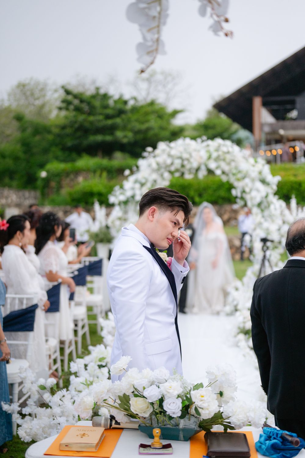 Emotional groom in white suit wiping tears during outdoor wedding ceremony with floral arch and white flower arrangements.