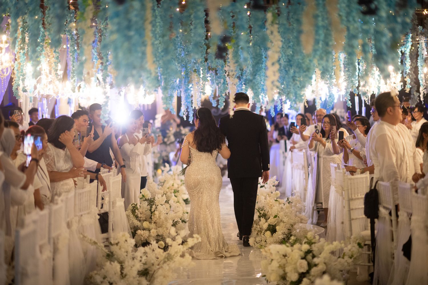 Elegant wedding ceremony with couple walking down floral aisle under hanging flowers, surrounded by guests in white attire.