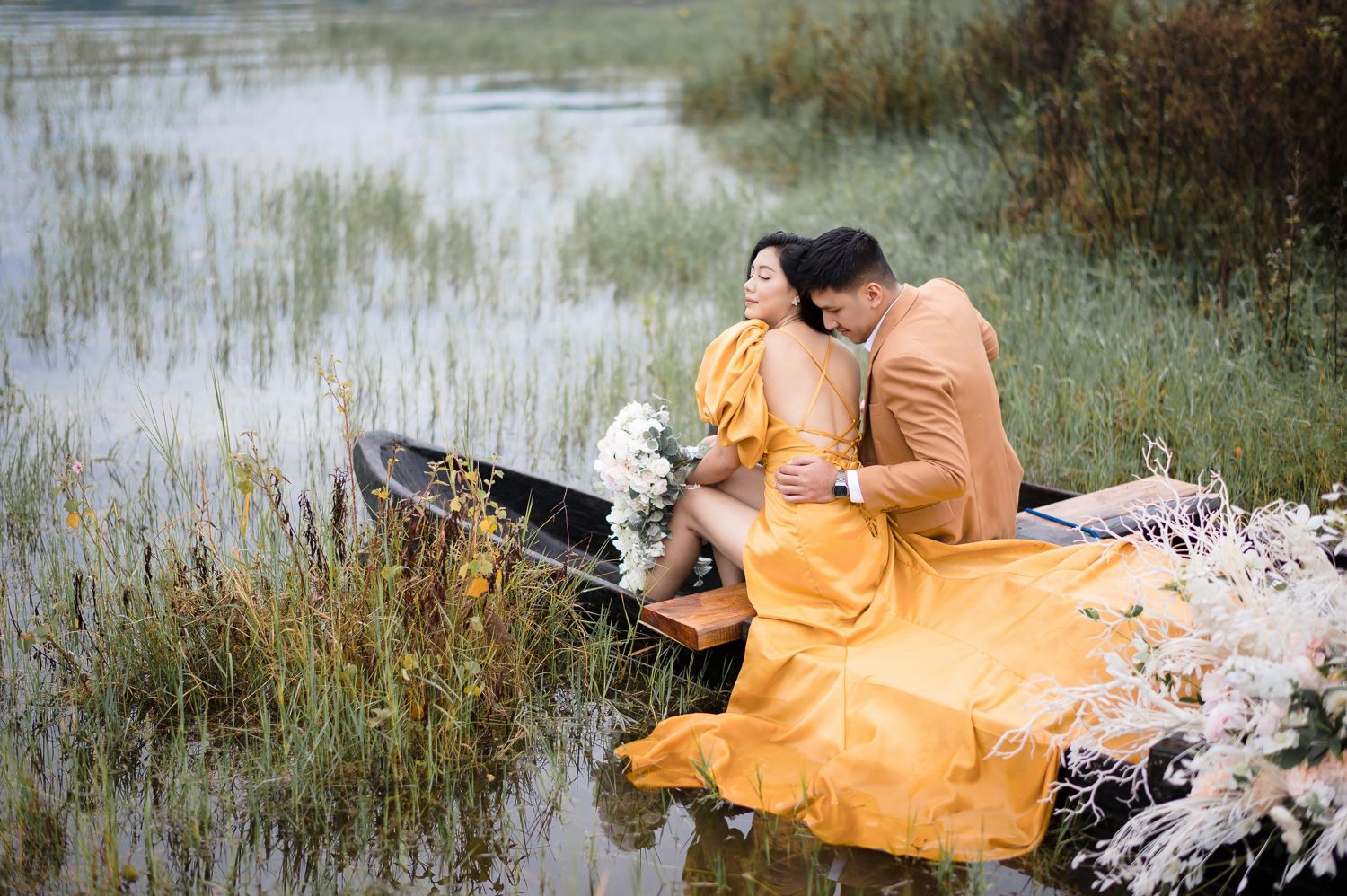 Couple in matching golden outfits sharing a romantic moment in a wooden boat surrounded by lush wetlands.