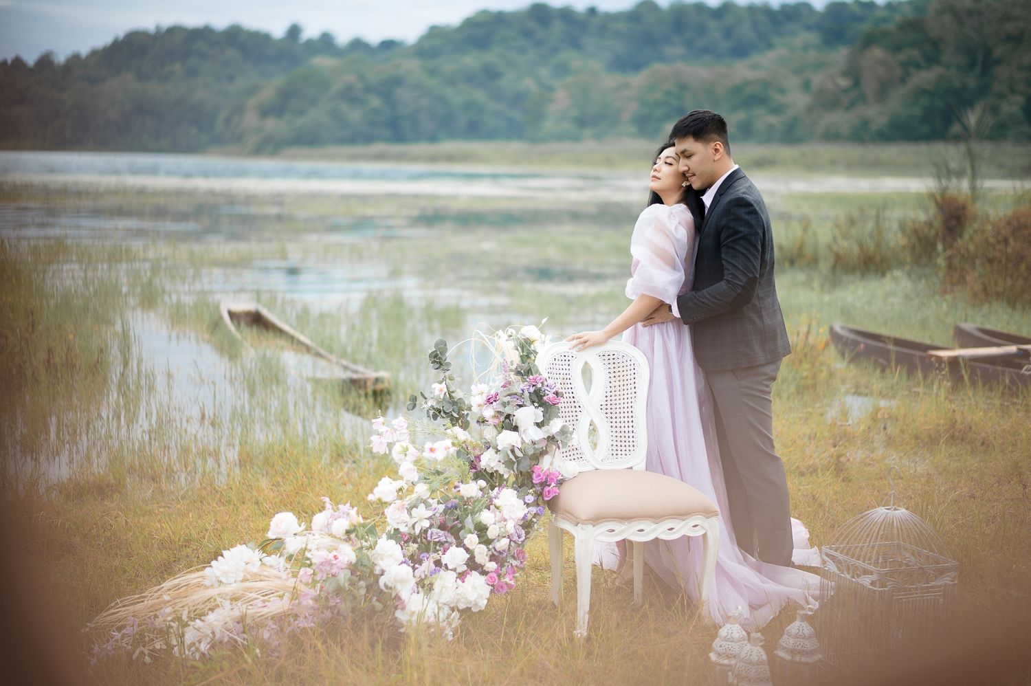 Romantic couple in wedding attire embracing by a serene lake with lush greenery and floral arrangements nearby.