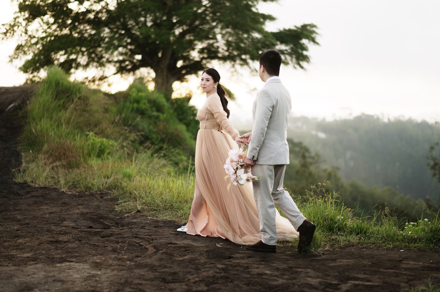 Couple in elegant attire walking on a scenic hilltop path surrounded by lush greenery at sunset.