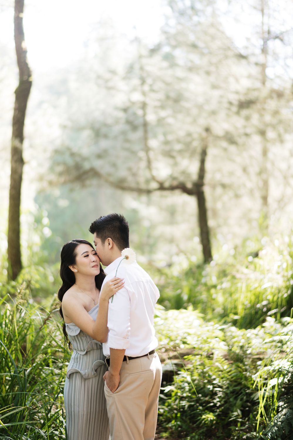 A couple shares a romantic embrace in a sunlit forest, the man kissing the woman's cheek among lush green foliage.