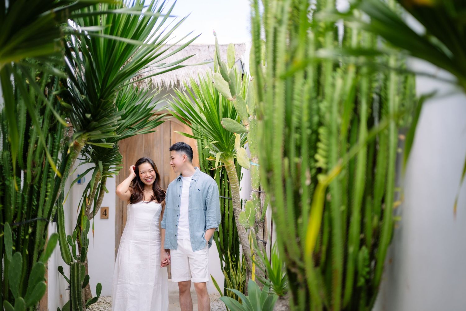 Couple walking through lush tropical garden path with cacti and palm plants, wearing casual white and blue outfits.