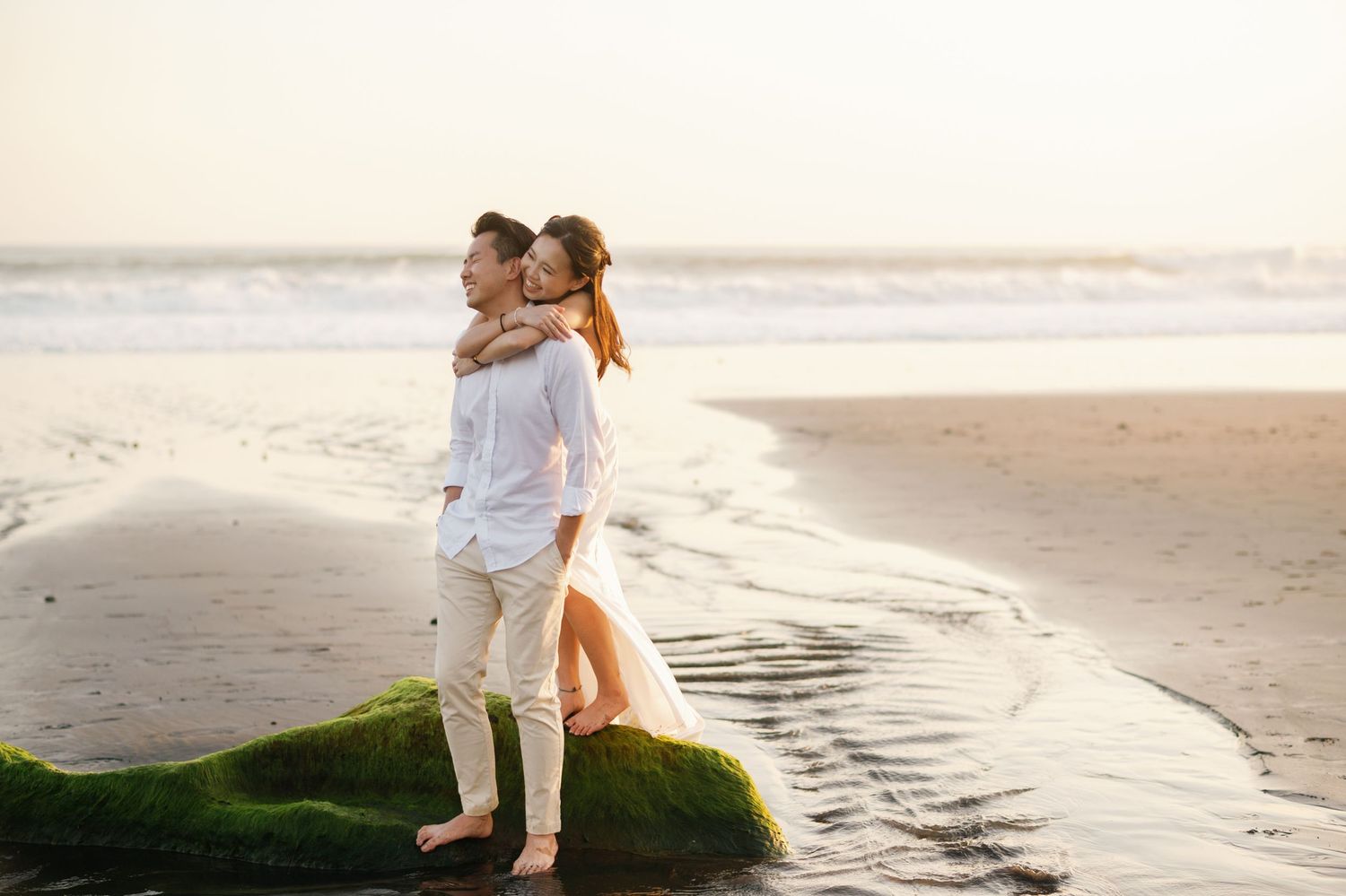 Romantic couple embracing on a mossy rock at the beach during golden hour, wearing white outfits.