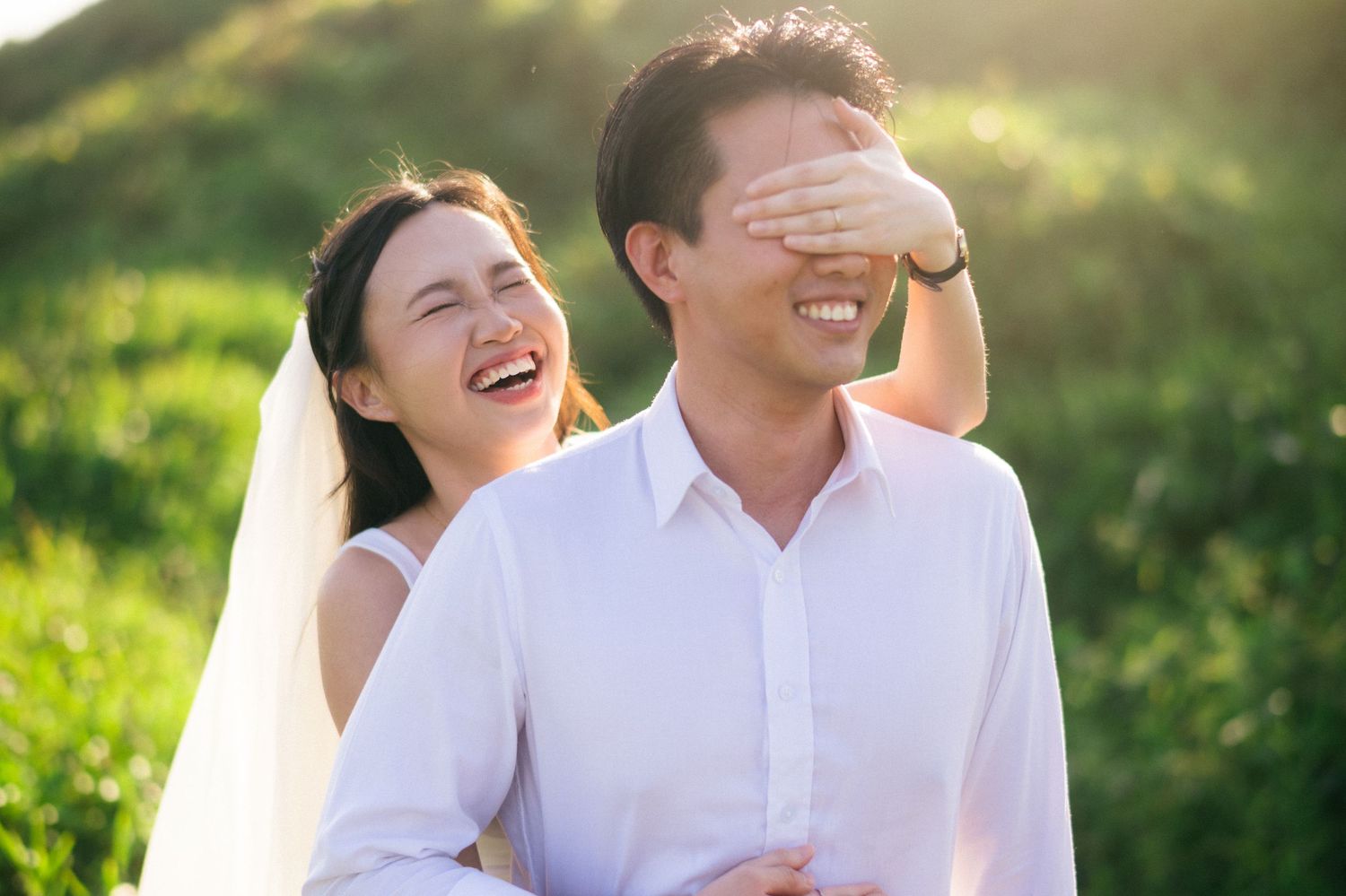 Happy Asian couple in wedding attire laughing outdoors, bride covering groom's eyes playfully in sunlit green field.
