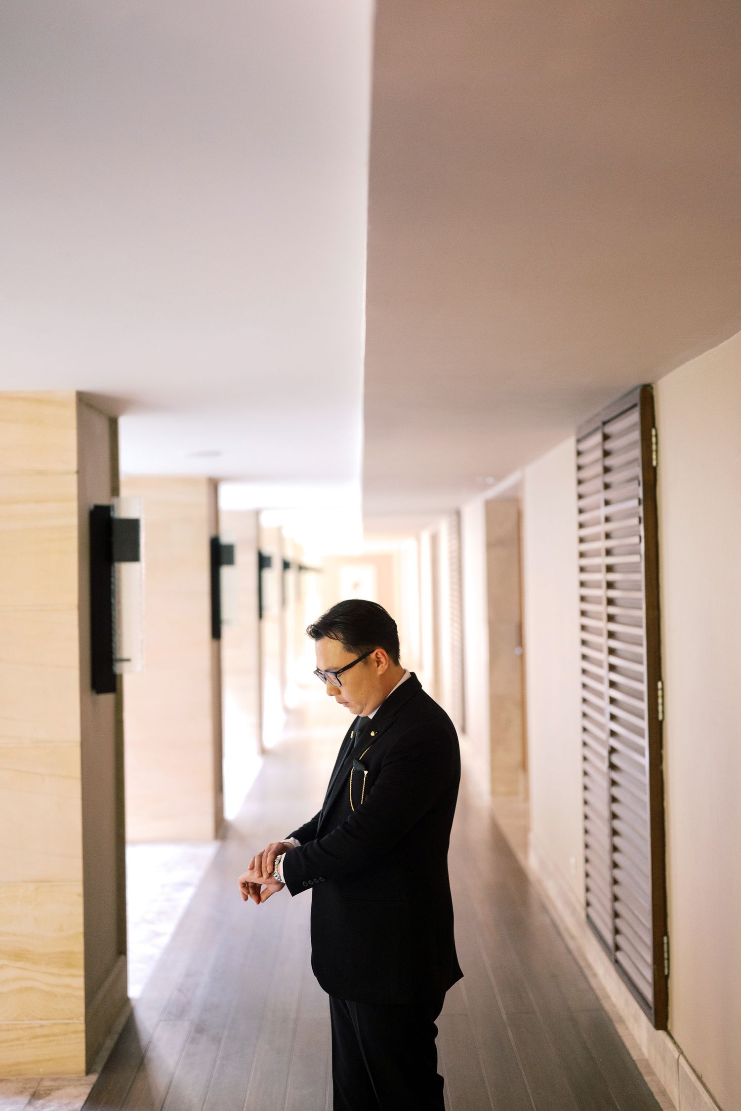 A well-dressed man in a black suit checks his watch while standing in a modern hotel corridor.