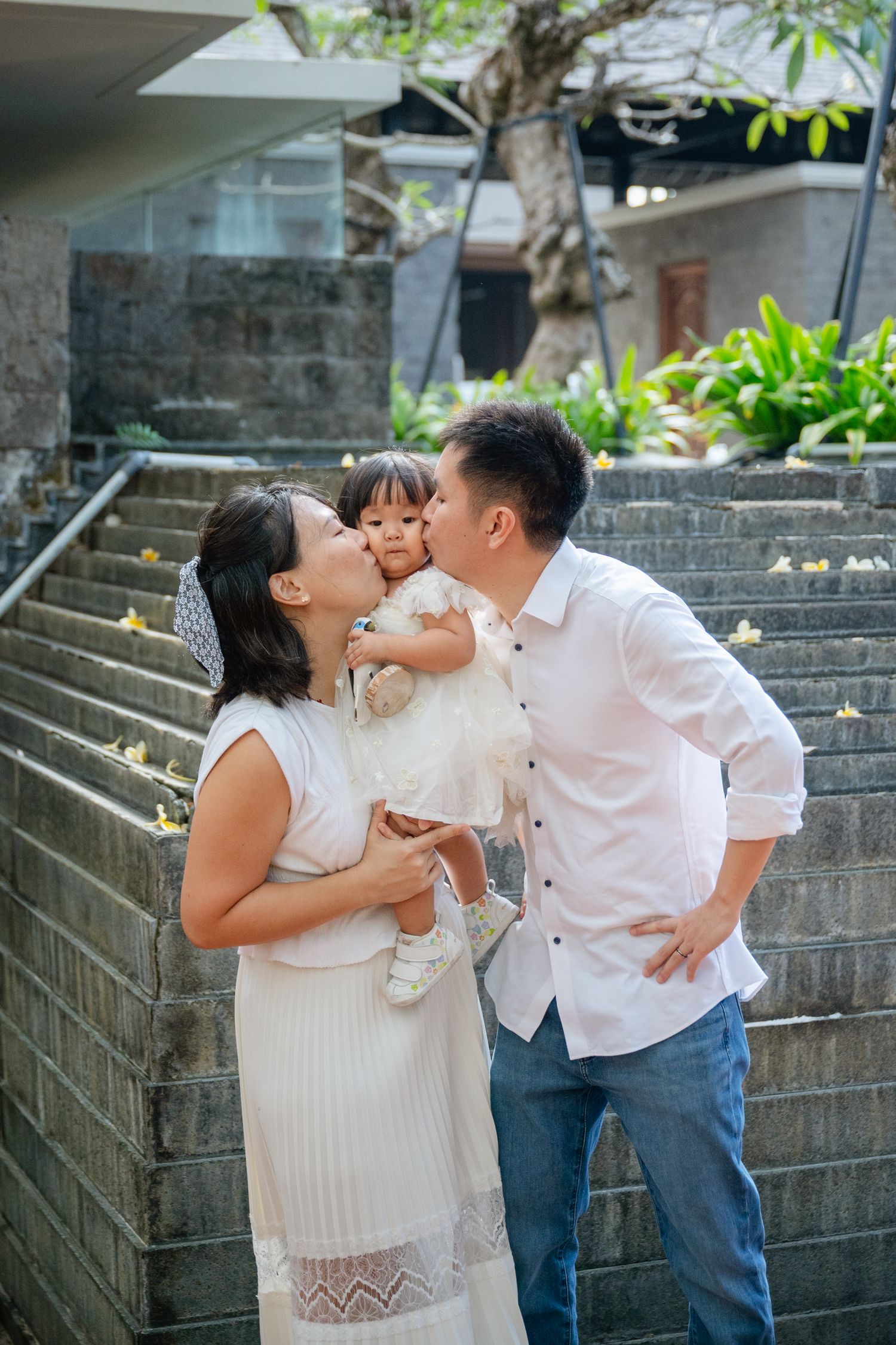 A couple in white outfits kisses their toddler daughter on the cheeks while standing on outdoor stone steps.