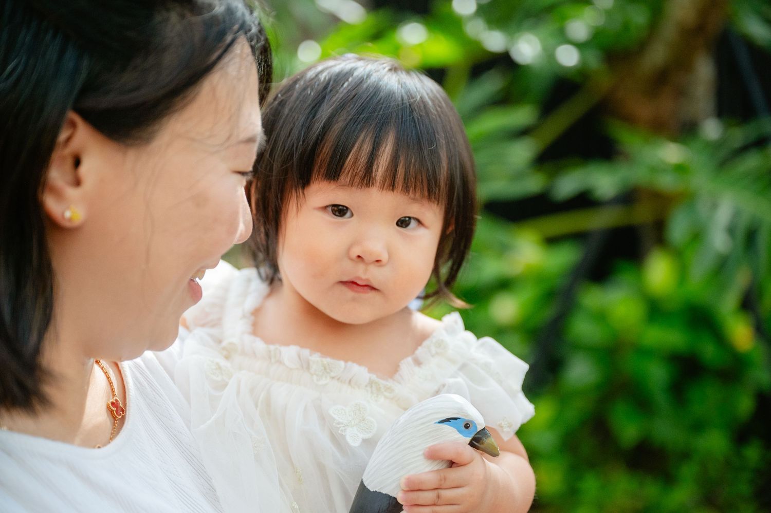 A young Asian toddler girl in a white dress being held by her mother outdoors in a lush green garden.