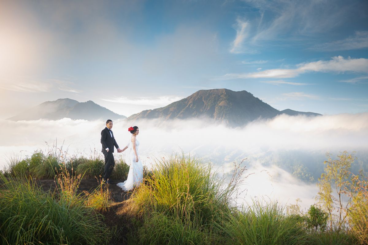 Couple in wedding attire standing on misty mountain peak above clouds, bride holding red bouquet with scenic valley backdrop.