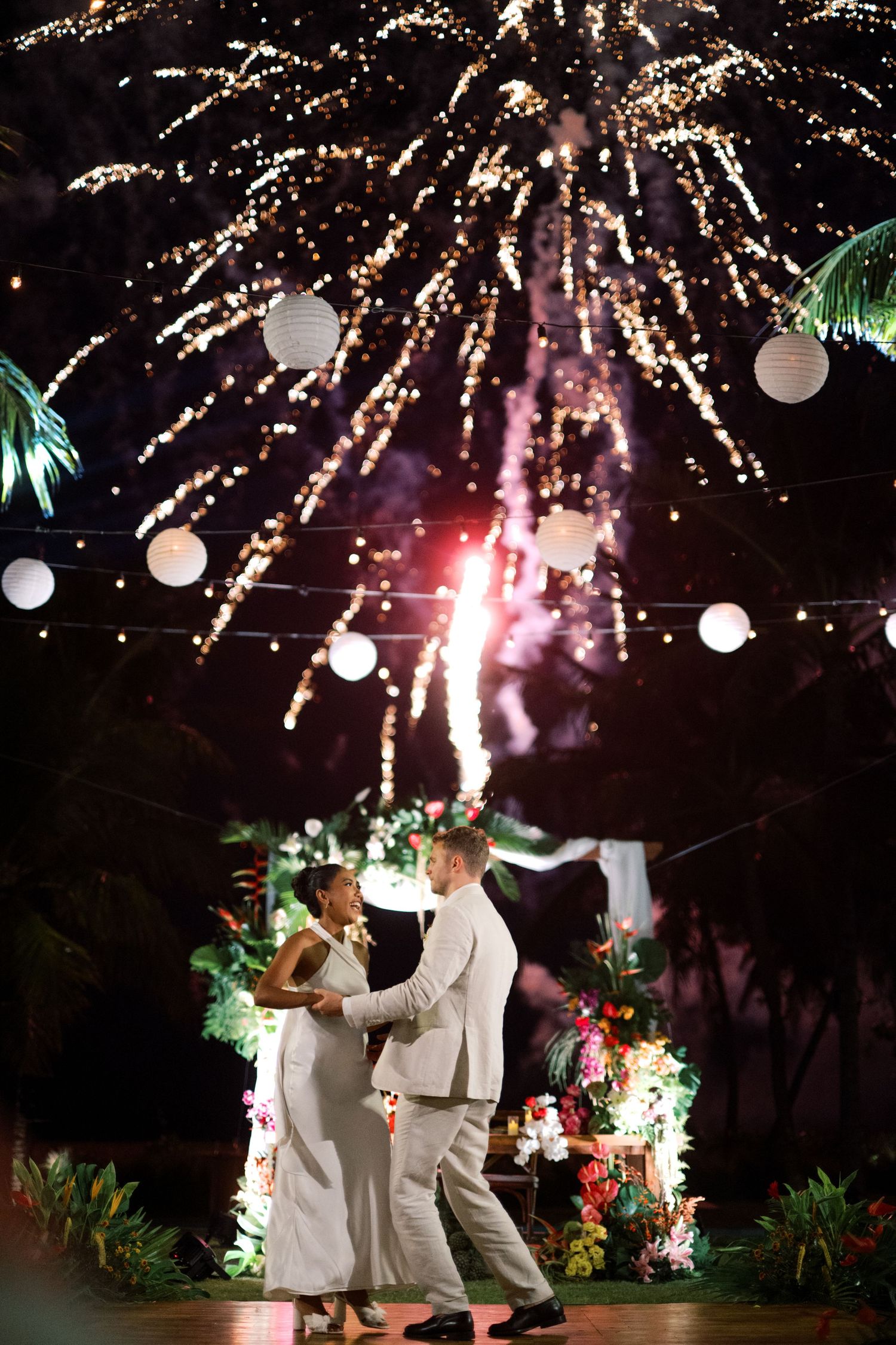 Couple shares first dance under spectacular fireworks display with white lanterns and tropical floral decor at outdoor wedding reception.