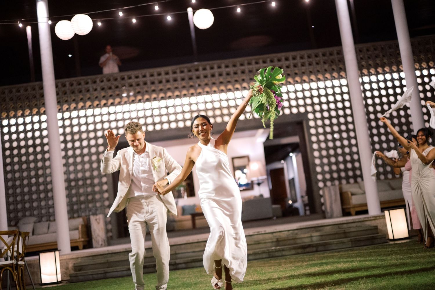 Joyful couple in white attire celebrating their wedding reception outdoors at night under string lights.