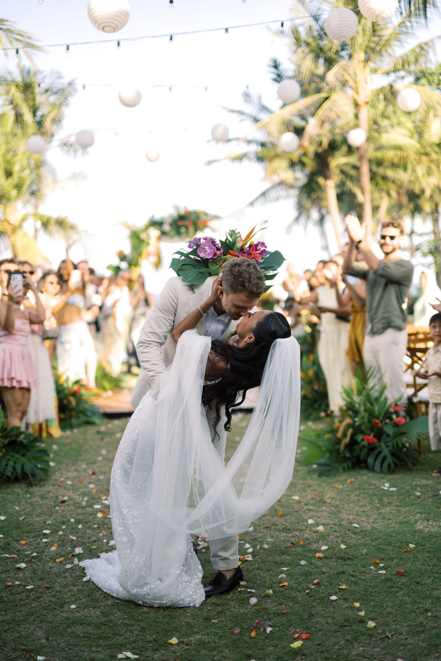 Bride and groom share a romantic dip kiss at outdoor tropical wedding ceremony with guests and palm trees.