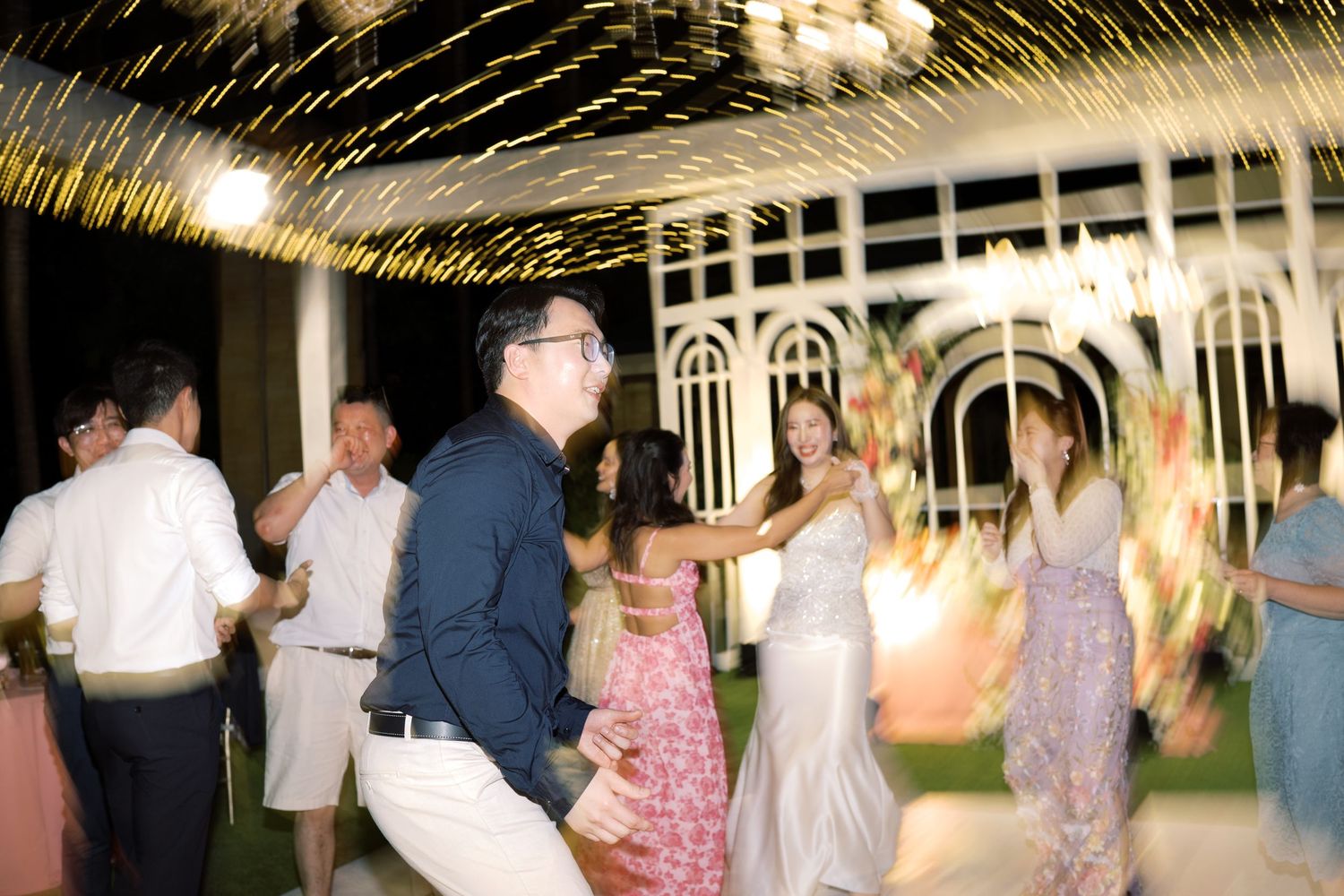 Guests dancing and celebrating outdoors at a wedding reception with sparkling lights and floral decorations at night.