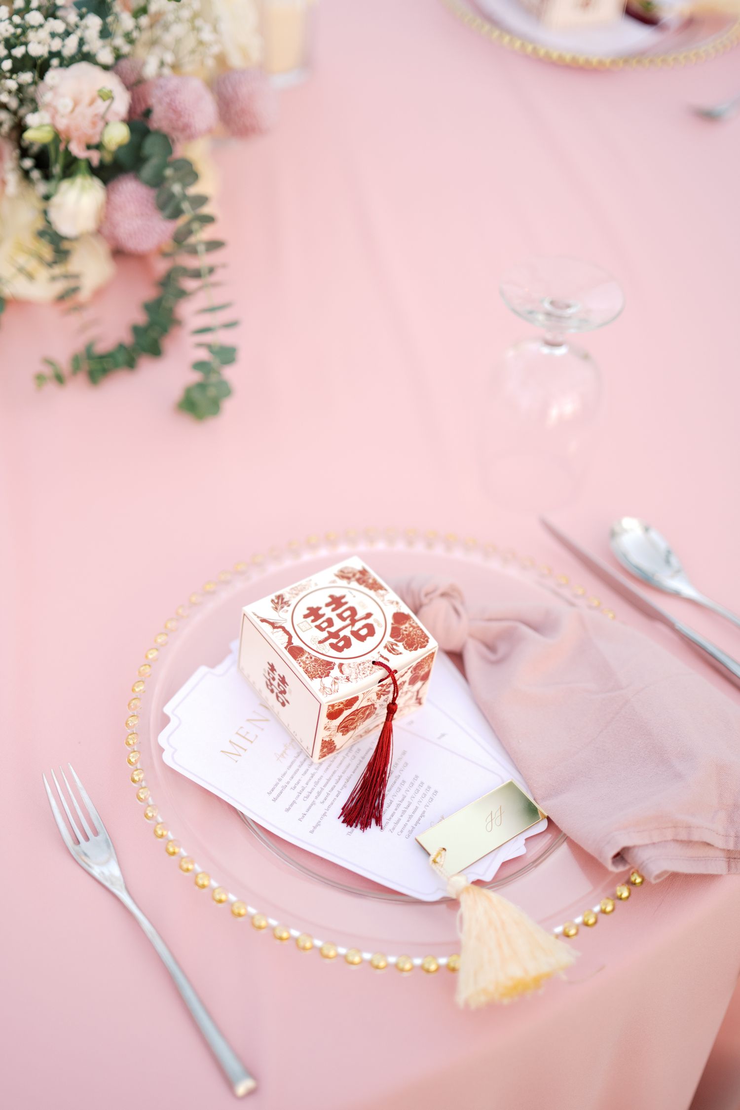 Elegant pink wedding table setting with gold-rimmed plate, floral favor box, tassel napkin ring, and fresh flowers.