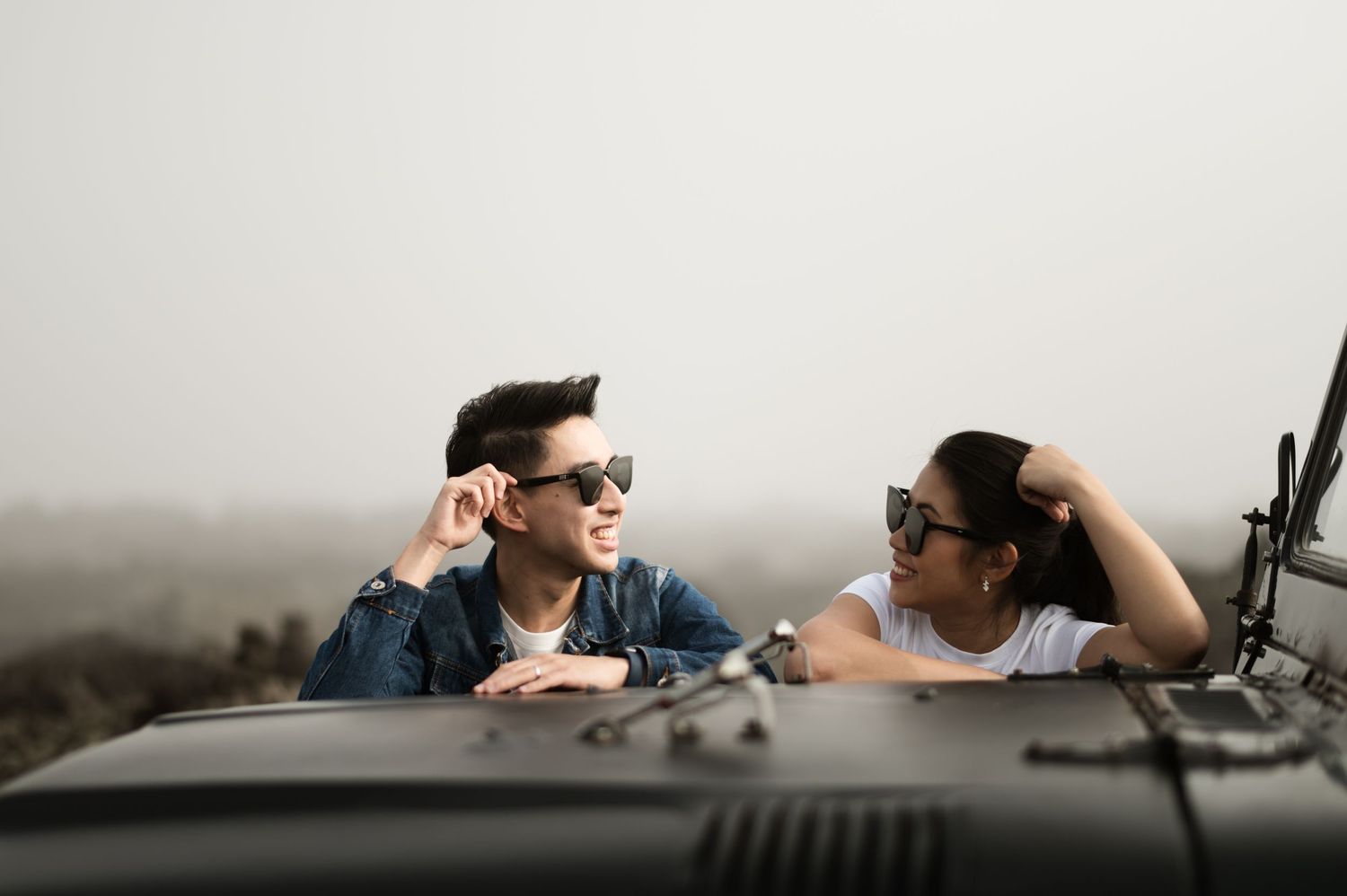 Two young people wearing sunglasses lean on a dark car hood, smiling and enjoying a scenic road trip together.