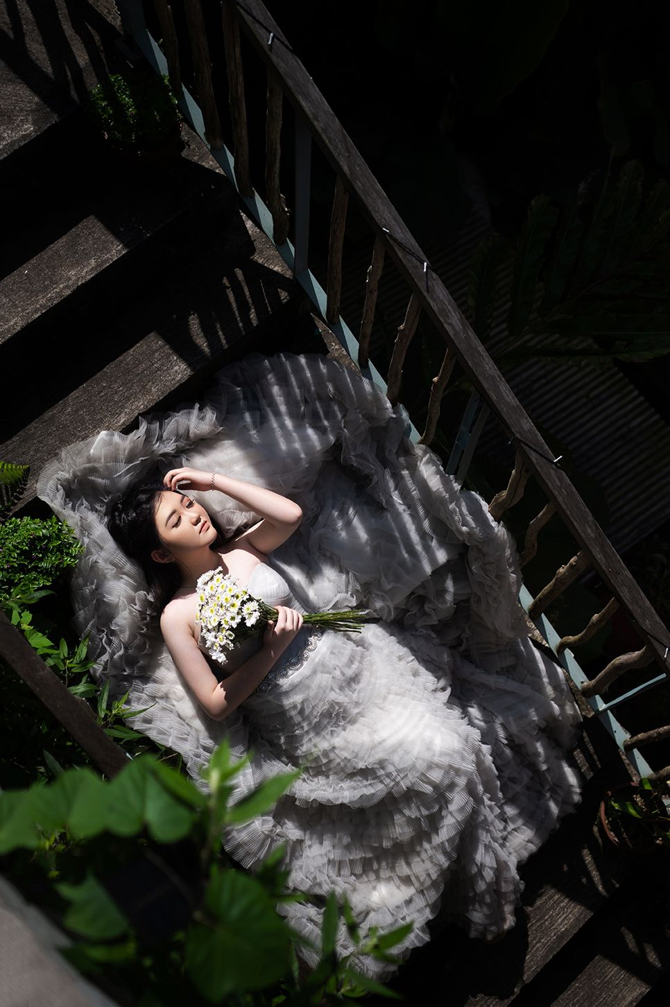 Bride in white wedding dress lying on wooden steps holding white flowers, viewed from above in dappled sunlight.