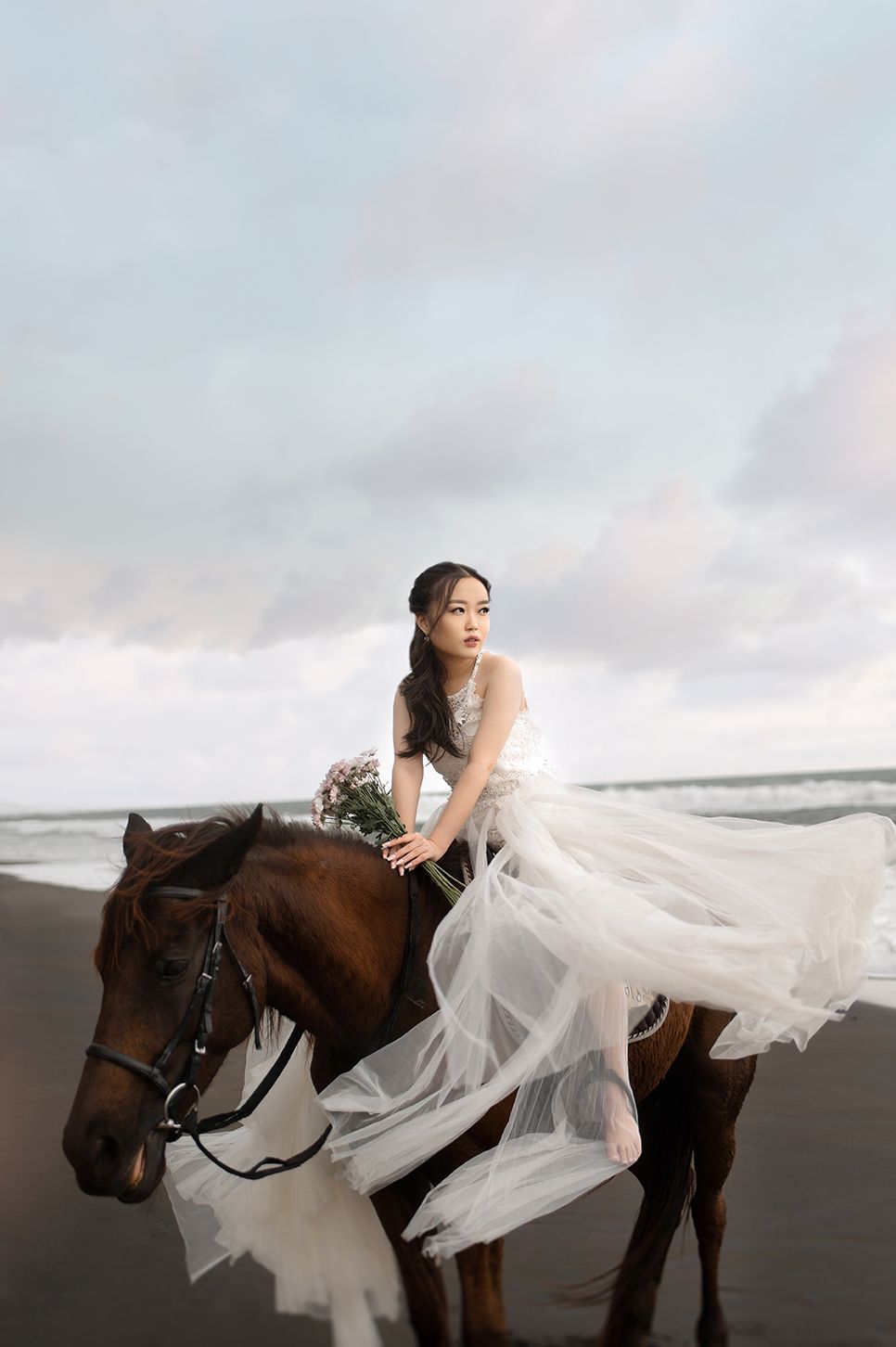 A bride in a flowing white tulle gown holds flowers while riding a brown horse on a dark sandy beach.