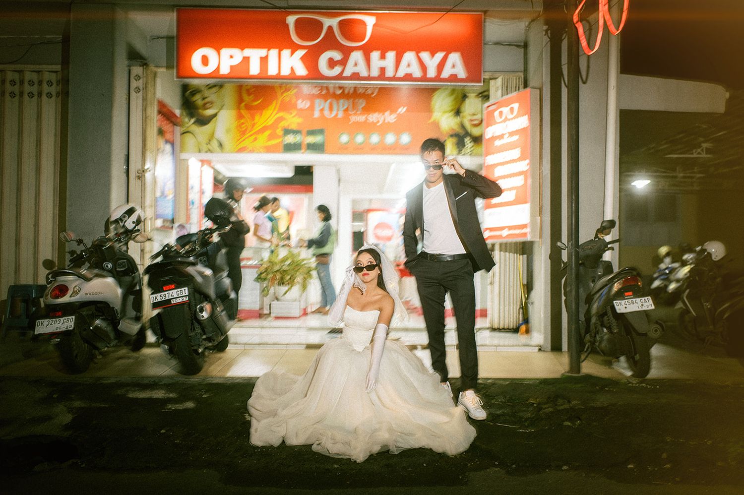 A bride in a white gown and groom in a black suit pose in front of Optik Cahaya store at night.
