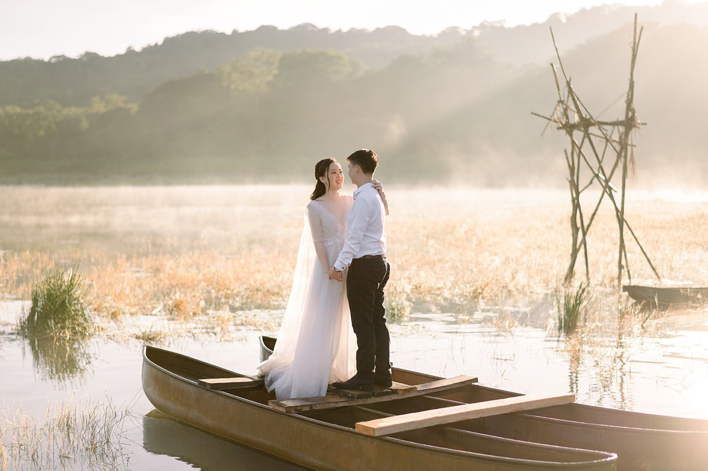 Couple in wedding attire standing romantically in a wooden boat on a misty lake at sunrise.