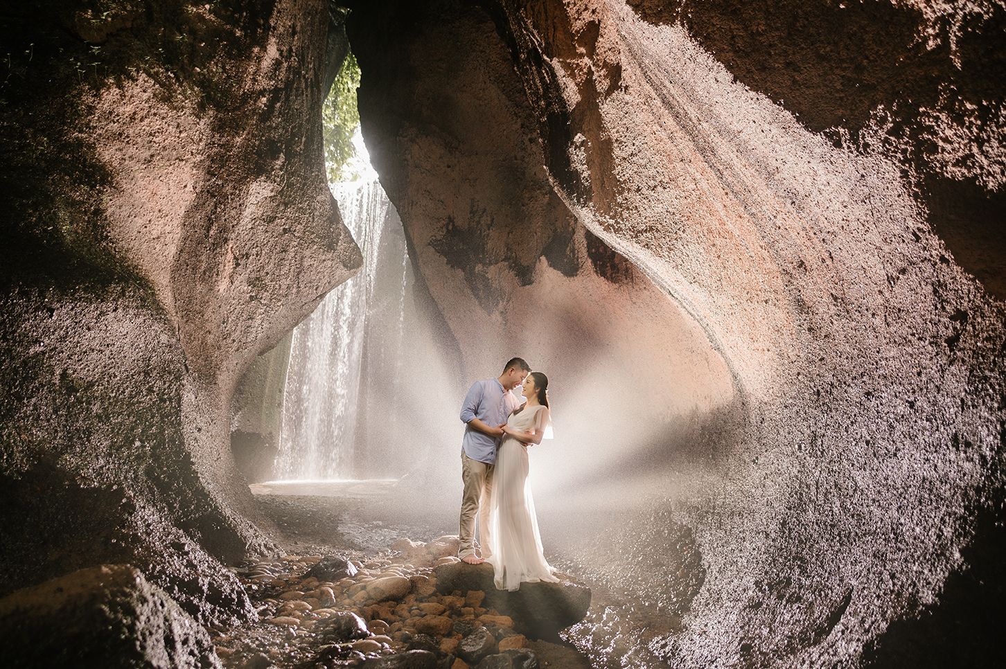 Couple embracing in a stunning cave with a waterfall and dramatic light rays, perfect for a romantic wedding photo.