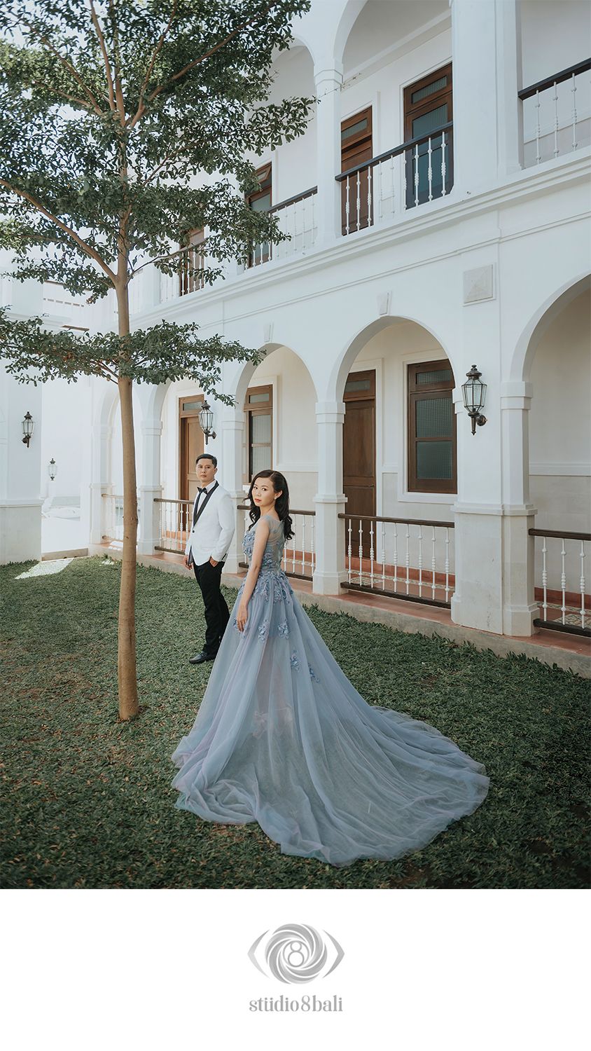 Couple poses in front of elegant white colonial building, woman in flowing blue-gray gown with long train, man in tuxedo.