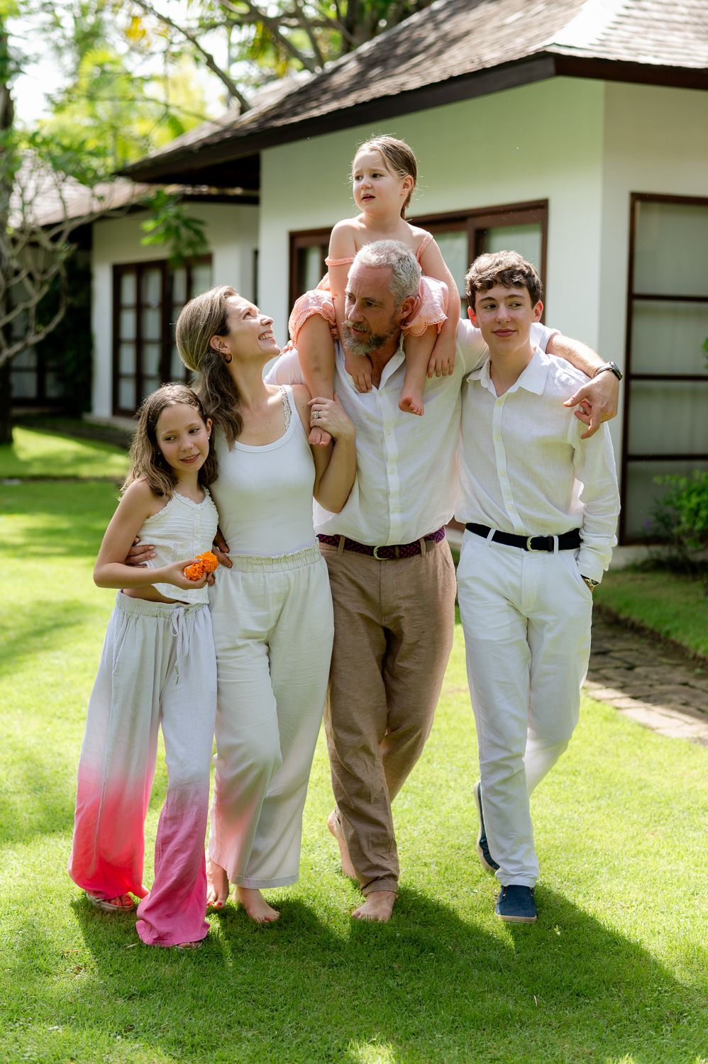 Happy family of five posing together on green lawn in front of white house, father carrying child on shoulders.
