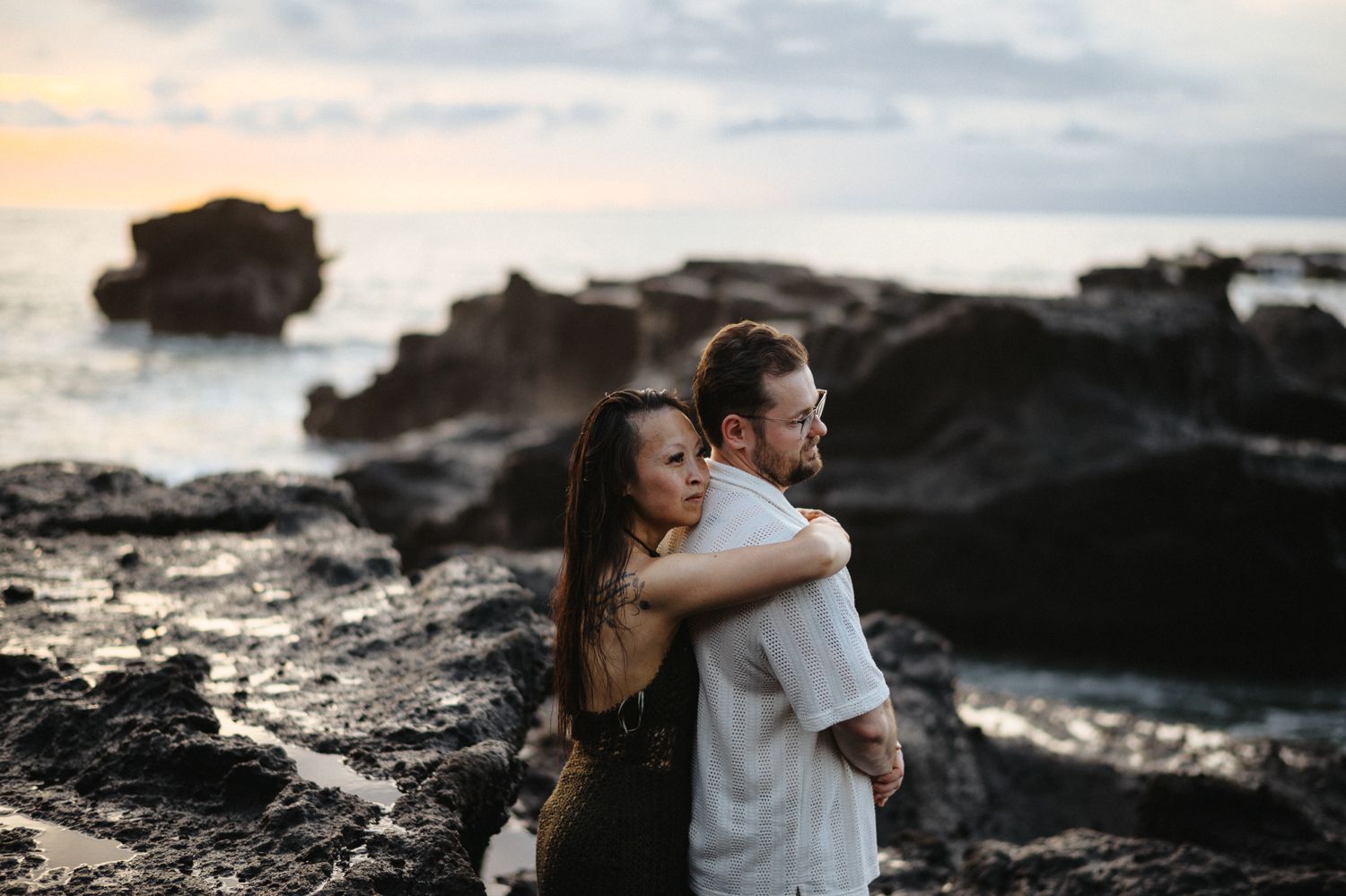 Couple embracing on rocky Hawaiian coastline at sunset, waves and dramatic rock formations in background.