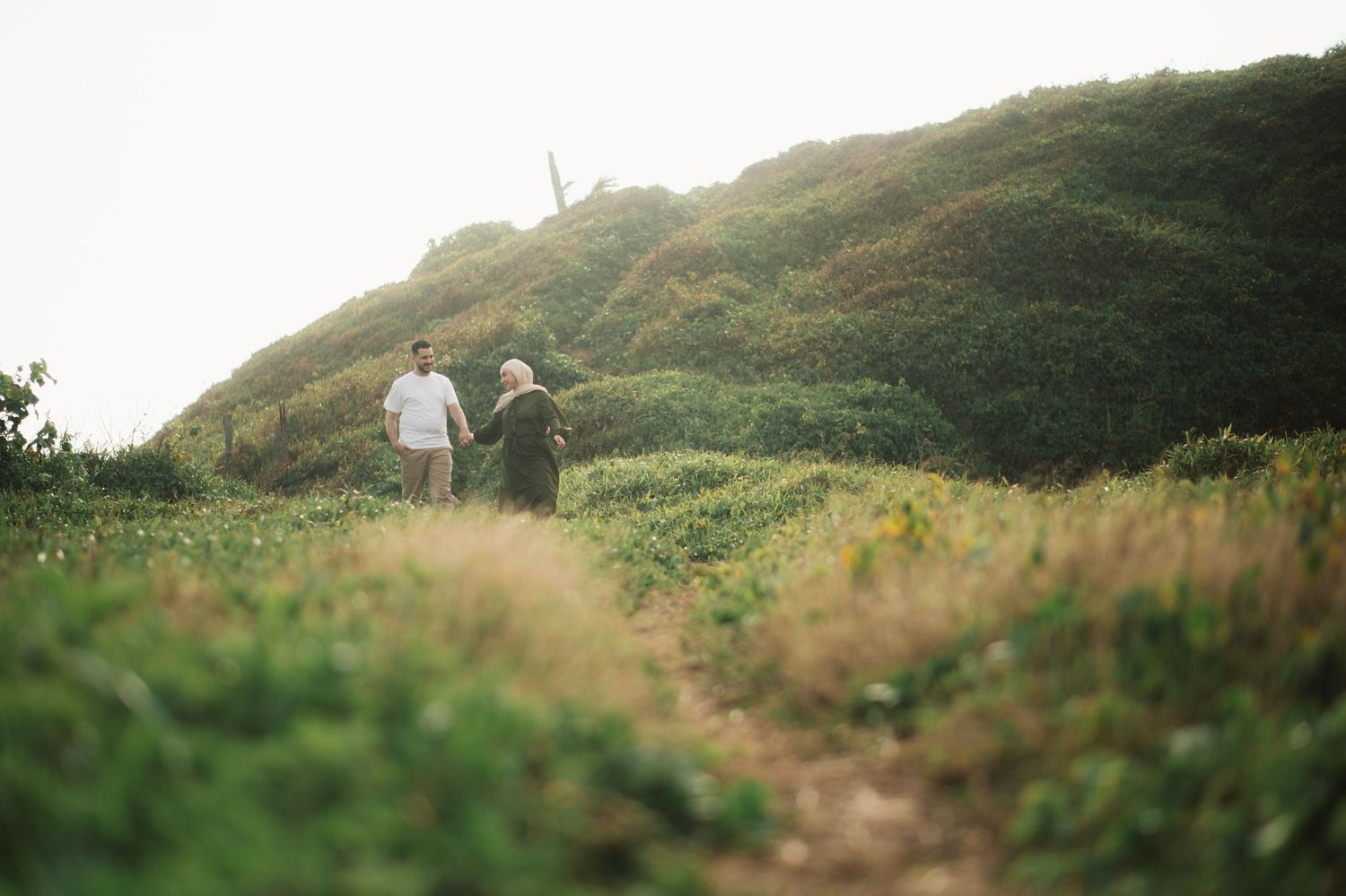 Two people hold hands while walking along a lush green hillside trail in a scenic outdoor landscape.