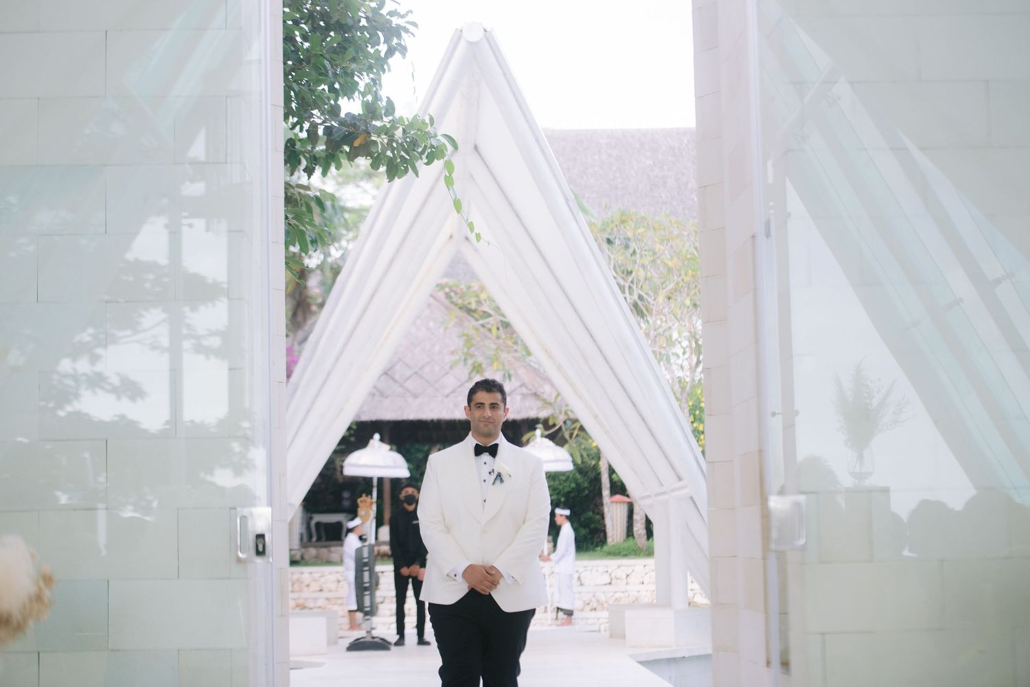 Groom in white tuxedo standing at elegant white draped wedding ceremony entrance doors.