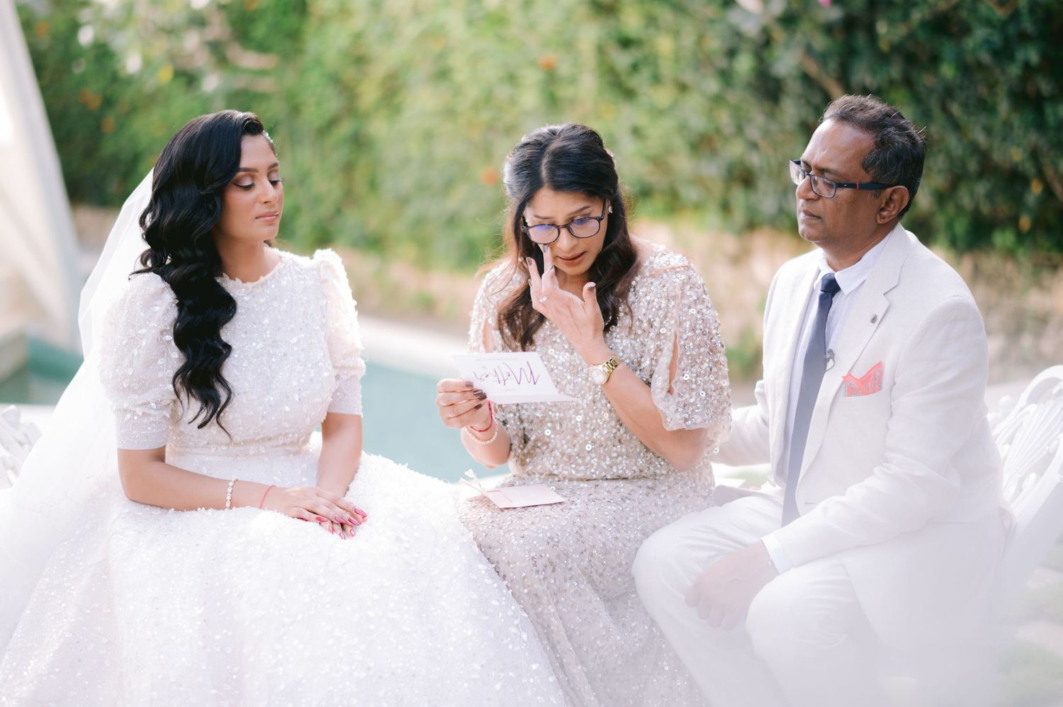 Bride in white gown with two guests reading a card at an elegant outdoor wedding ceremony.