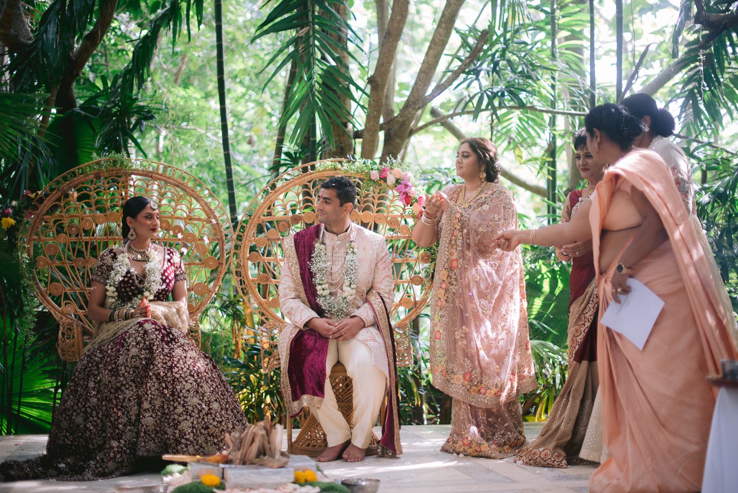 Couple in traditional Indian wedding attire seated on ornate chairs outdoors, surrounded by lush greenery and guests in elegant sarees.