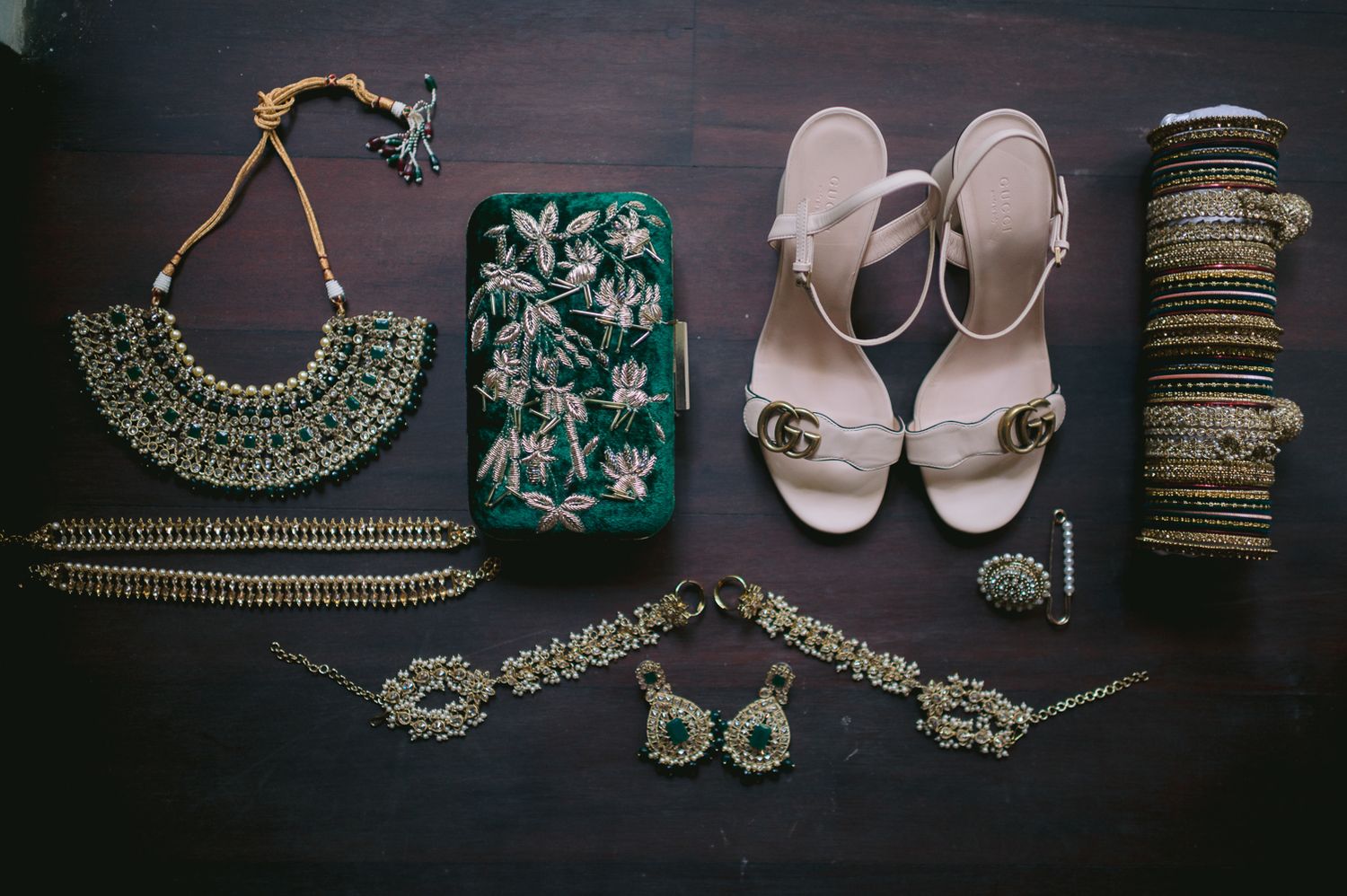 Flat lay of Indian bridal accessories including gold jewelry, green embroidered clutch, nude Gucci heels, and bangles.