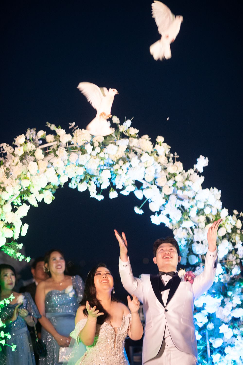 Newlywed couple releasing white doves at night wedding ceremony under floral arch with glowing white flowers.